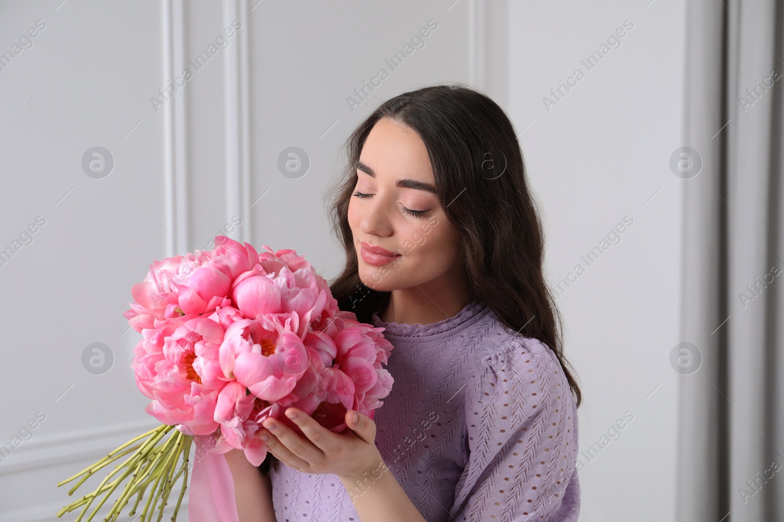 Photo of Beautiful young woman with bouquet of pink peonies near white wall indoors