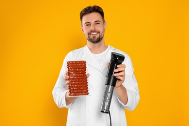 Photo of Smiling chef holding sous vide cooker and sausages in vacuum pack on orange background