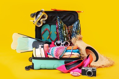 Open suitcase with clothes, beach accessories and shoes on yellow background. Summer vacation