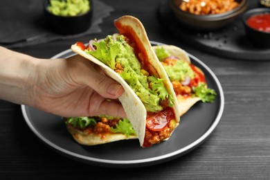 Photo of Woman holding taco with guacamole, meat and vegetables at wooden table, closeup