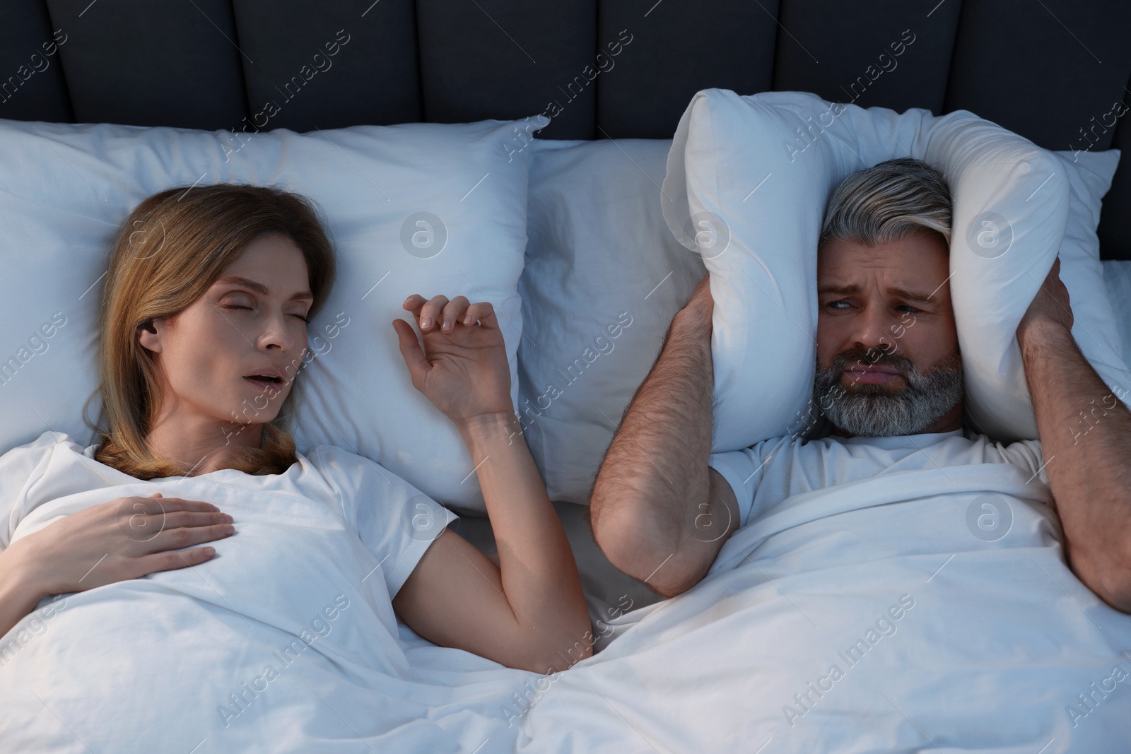 Photo of Irritated man covering his ears with pillows in bed at home. Problem with snoring wife