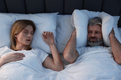 Photo of Irritated man covering his ears with pillows in bed at home. Problem with snoring wife