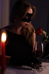 Elegant woman in black eye mask with rose and glass of wine at table indoors in evening