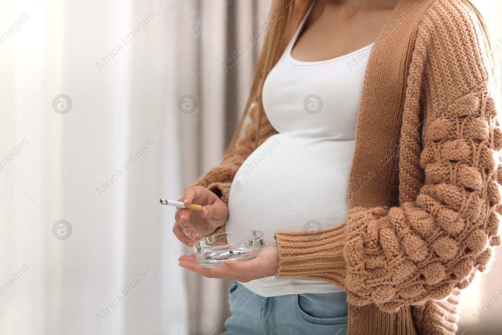 Photo of Young pregnant woman smoking cigarette at home, closeup. Harm to unborn baby