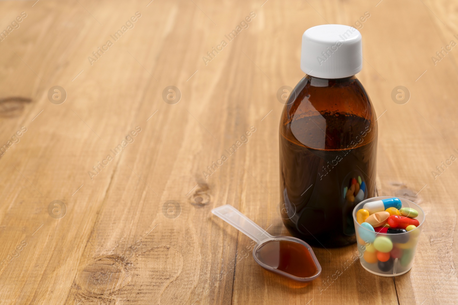 Photo of Bottle of syrup, dosing spoon and measuring cup with pills on wooden table, space for text. Cold medicine