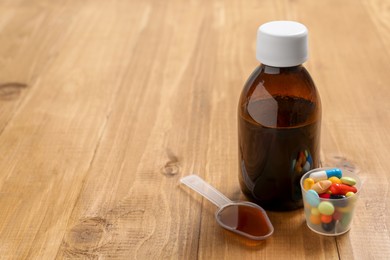 Photo of Bottle of syrup, dosing spoon and measuring cup with pills on wooden table, space for text. Cold medicine