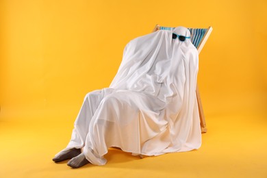 Person in ghost costume and sunglasses relaxing on deckchair against yellow background