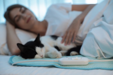 Photo of Young woman and cat in bed with electric heating pad, focus on cable