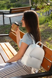 Photo of Beautiful young woman with stylish backpack and cup of coffee on bench in park