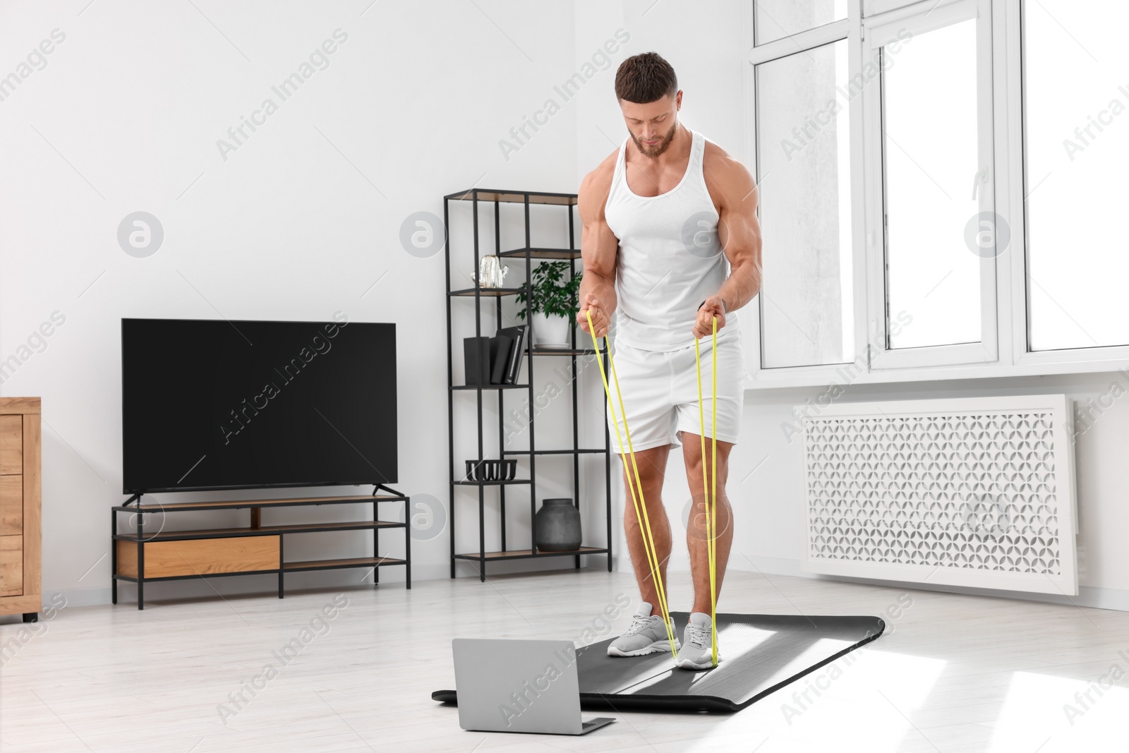 Photo of Muscular man doing exercise with elastic resistance band near laptop on mat at home