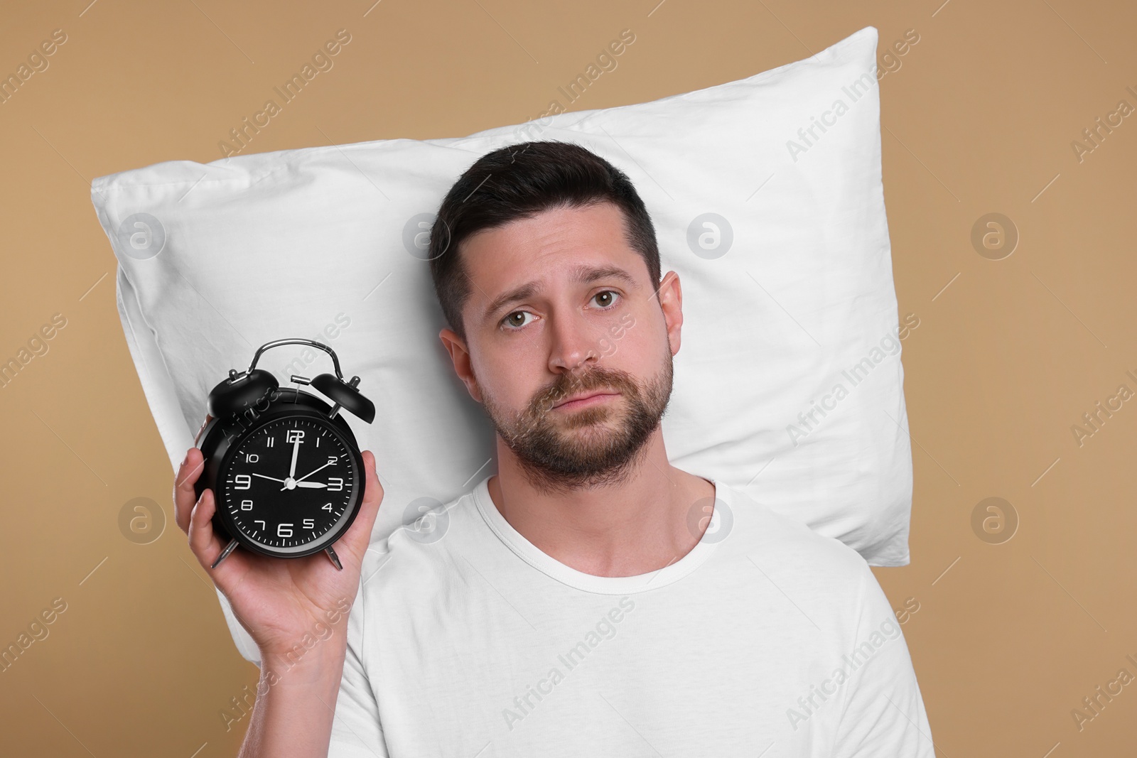 Photo of Sleepy man with pillow and alarm clock on beige background. Insomnia problem