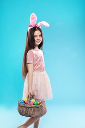 Little girl in bunny ears headband holding basket with Easter eggs on color background