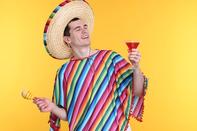 Photo of Young man in Mexican sombrero hat and poncho with cocktail and maracas on yellow background