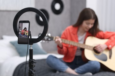 Photo of Teenage blogger playing guitar while streaming at home, focus on smartphone