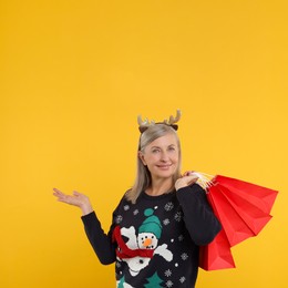 Happy senior woman in Christmas sweater and deer headband with shopping bags on orange background