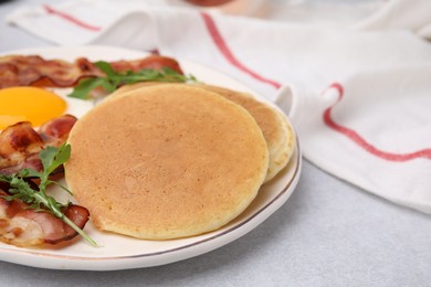 Photo of Plate with tasty pancakes, fried egg, arugula and bacon on light grey textured table, closeup. Space for text