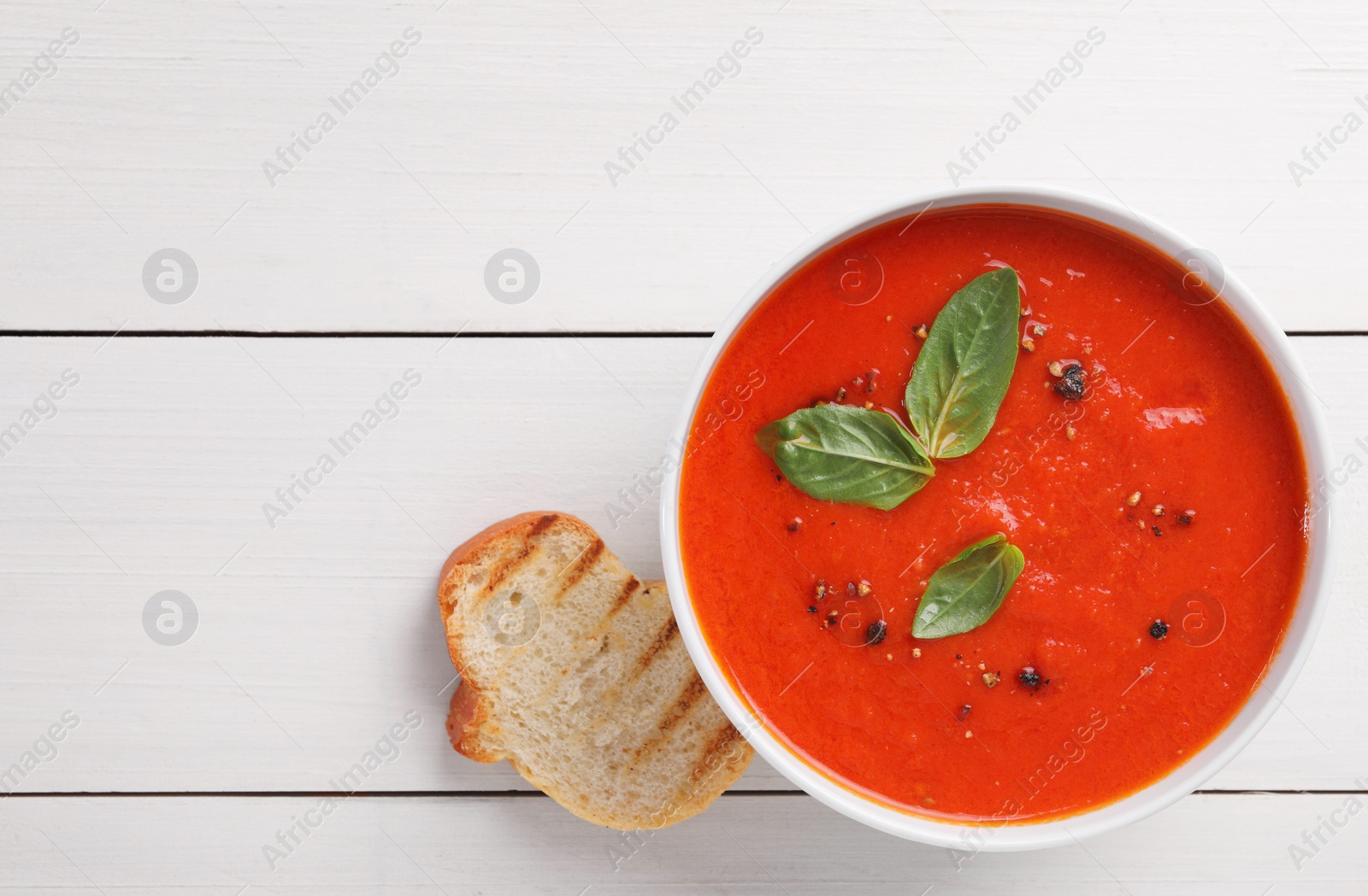 Photo of Delicious tomato cream soup in bowl and piece of grilled bread on white wooden table, flat lay. Space for text