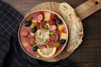 Photo of Meat solyanka soup with thin dry smoked sausages in bowl and bread slices on wooden table, flat lay