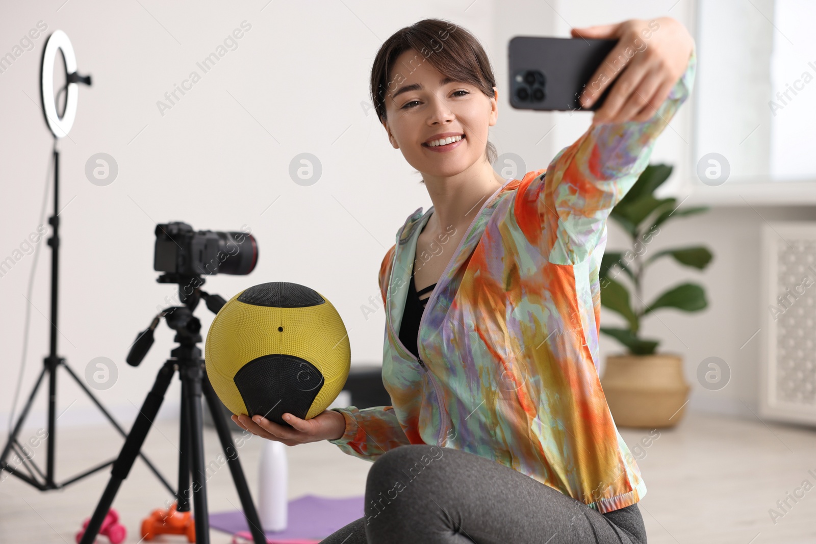 Photo of Happy sports blogger holding medicine ball while streaming online fitness lesson with smartphone at home