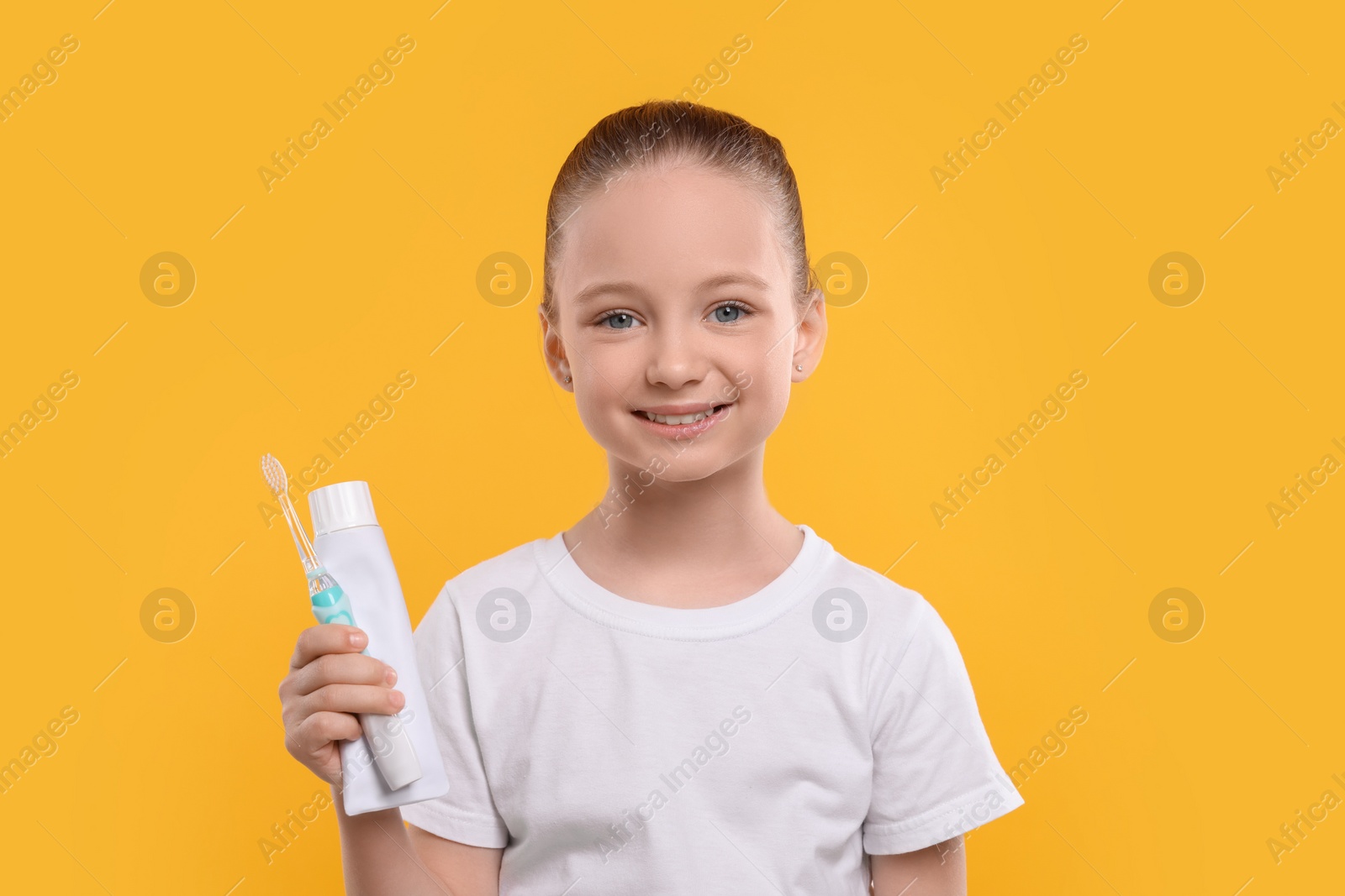 Photo of Happy girl holding electric toothbrush and tube of toothpaste on yellow background