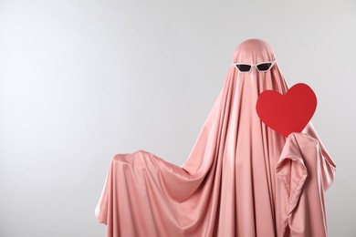 Photo of Cute ghost. Woman in pink sheet with sunglasses and red heart on light grey background, space for text