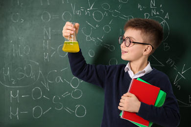 Photo of Schoolboy holding flask and notebooks near chalkboard with chemical formulas