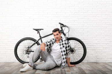 Photo of Handsome young man with modern bicycle near white brick wall indoors