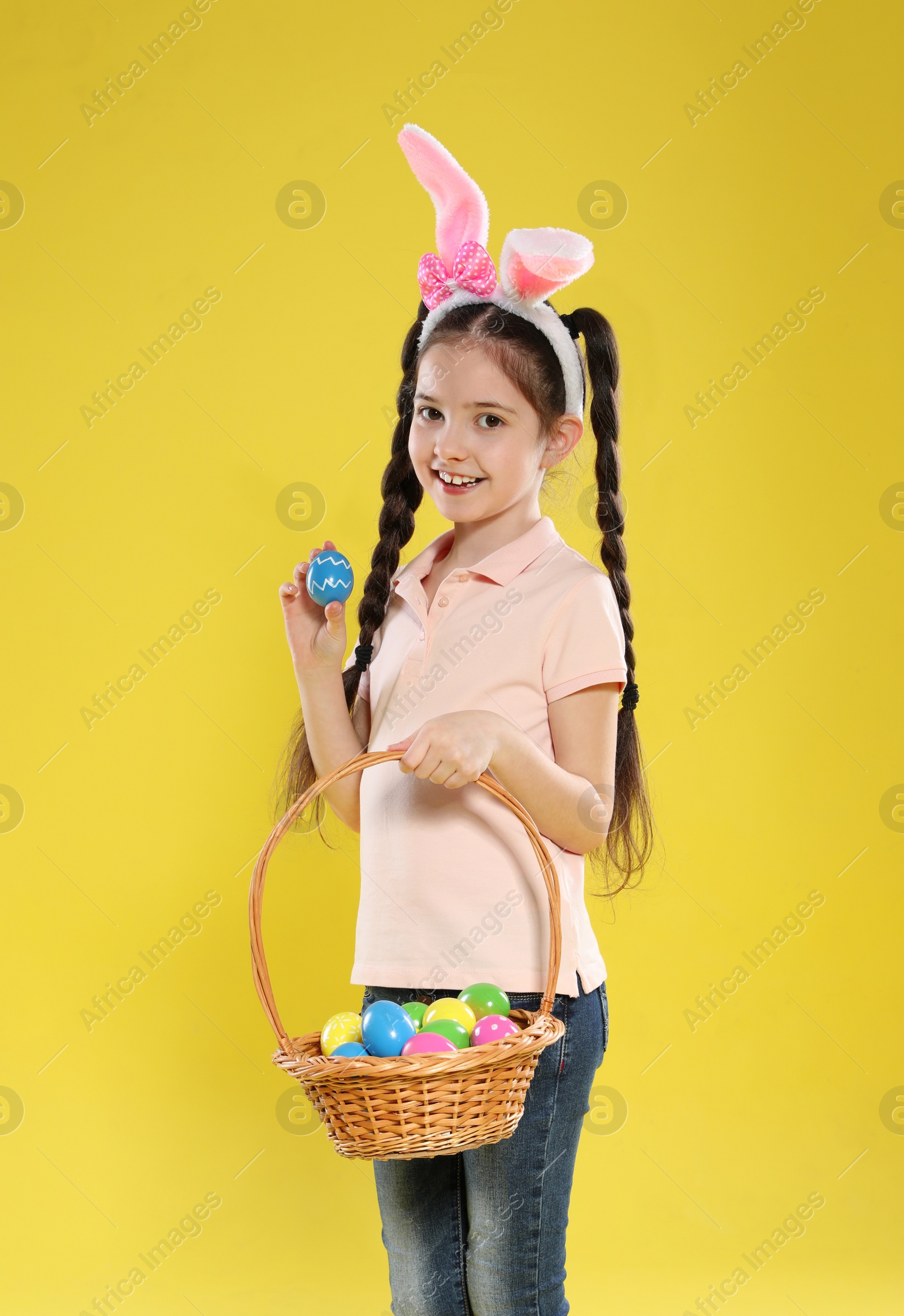 Photo of Little girl in bunny ears headband holding basket with Easter eggs on color background