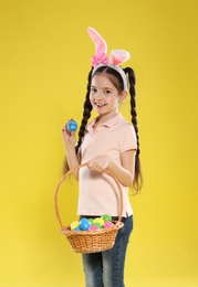 Little girl in bunny ears headband holding basket with Easter eggs on color background