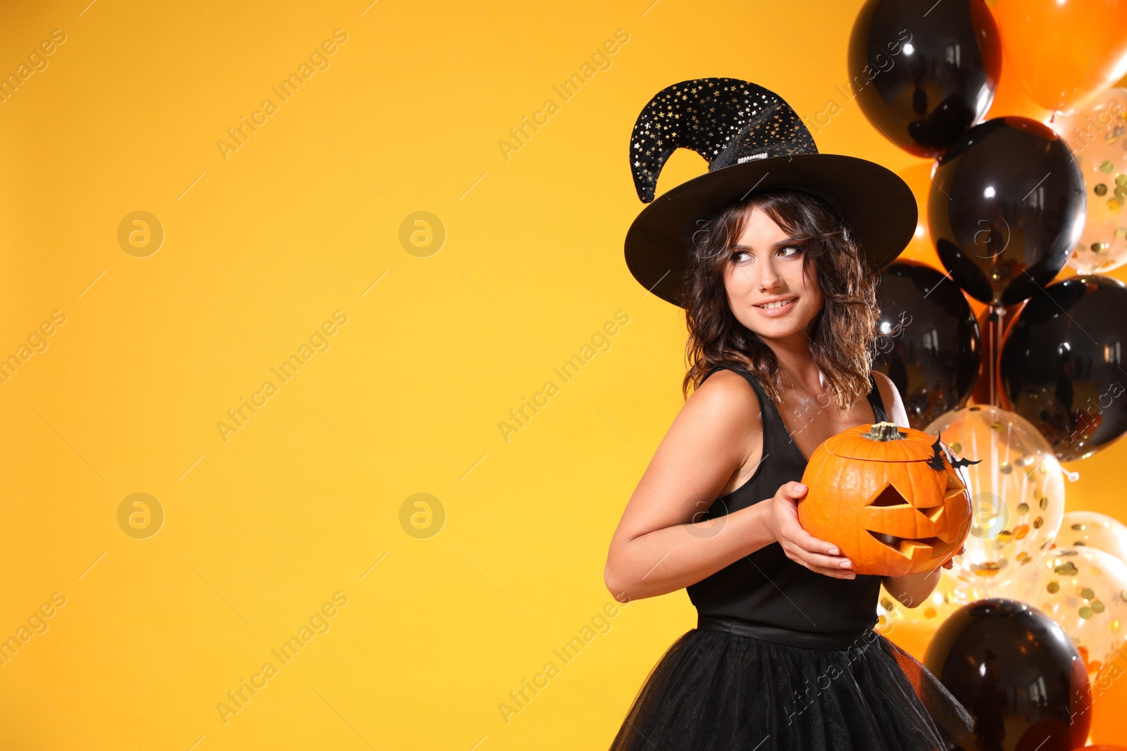 Photo of Beautiful woman in witch costume with balloons and jack o'lantern on yellow background, space for text. Halloween party