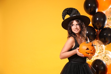 Beautiful woman in witch costume with balloons and jack o'lantern on yellow background, space for text. Halloween party