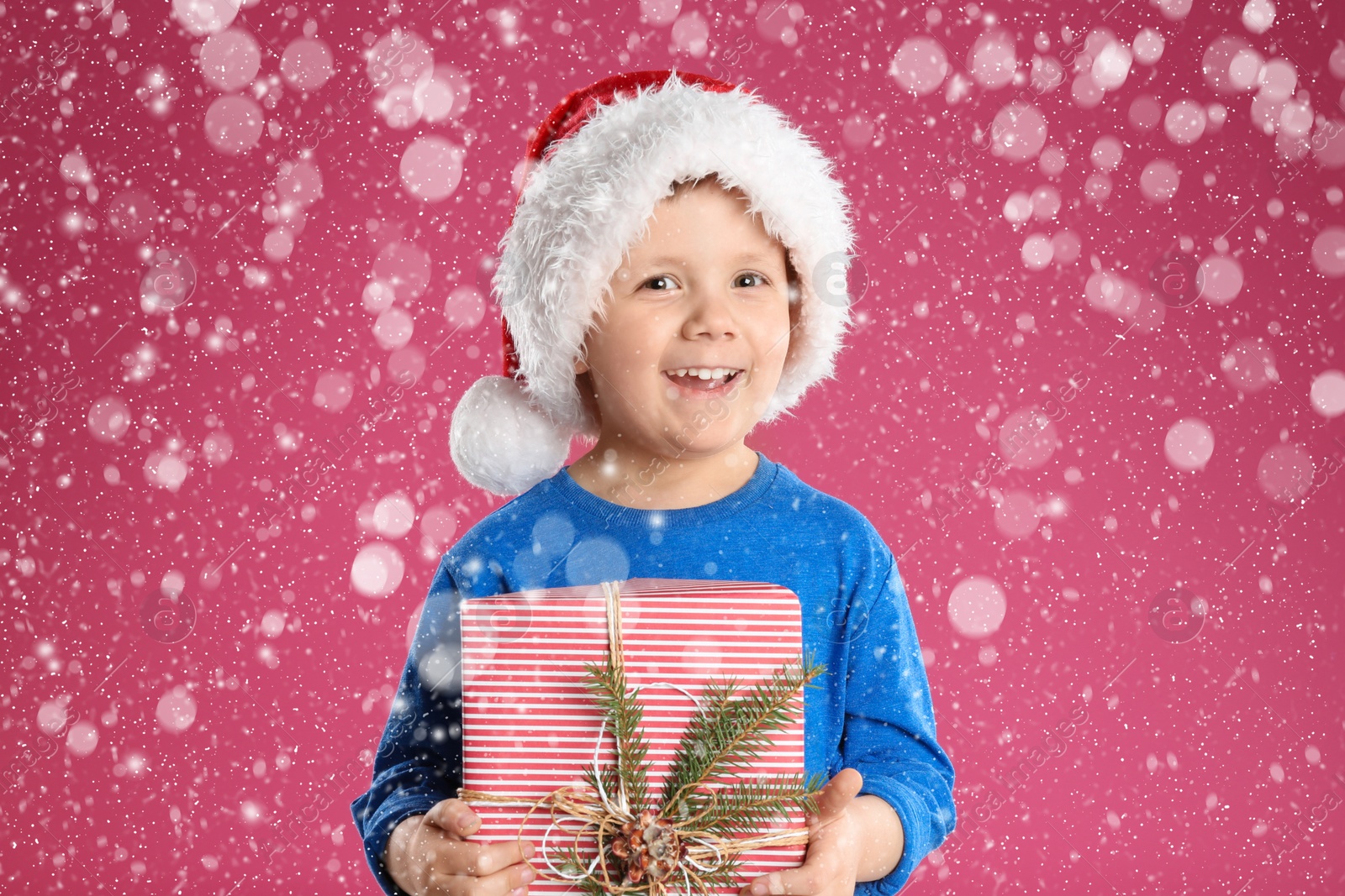 Image of Cute child in Santa hat with Christmas gift under snowfall on pink background