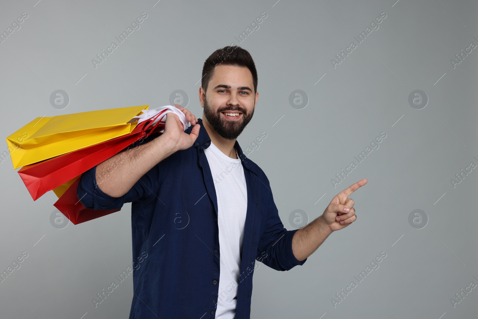 Photo of Happy man with many paper shopping bags pointing at something on grey background. Space for text