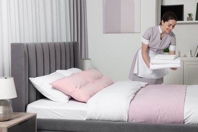 Photo of Young maid with stack of towels near bed in hotel room