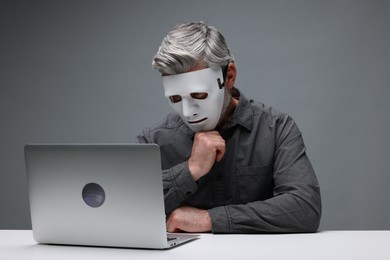 Man in mask working with laptop at white table against grey background