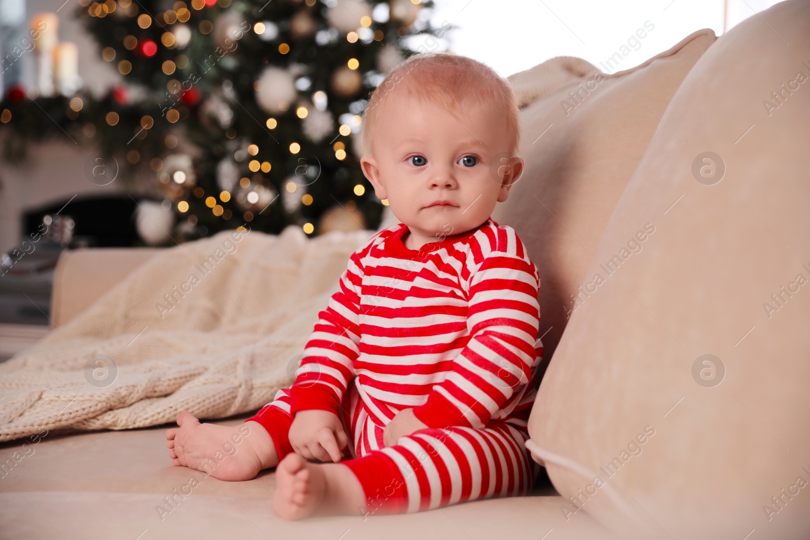 Photo of Cute baby in bright Christmas pajamas sitting on sofa at home