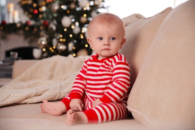 Cute baby in bright Christmas pajamas sitting on sofa at home