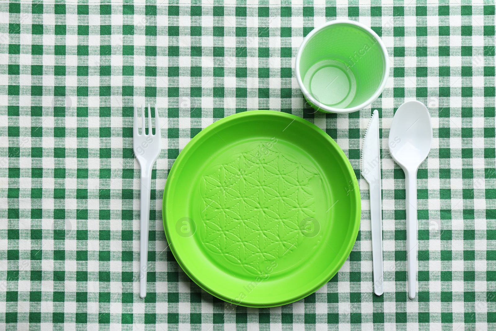 Photo of Composition with plastic dishware on checkered tablecloth, top view. Picnic table setting