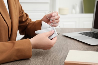 Woman wiping her glasses with microfiber cloth at wooden table in office, closeup