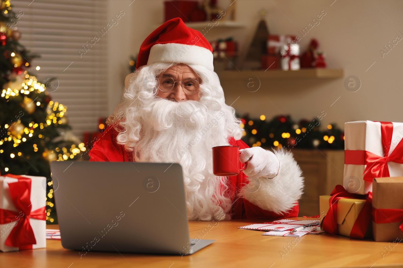 Photo of Santa Claus holding cup of drink using laptop at table with Christmas gifts in room