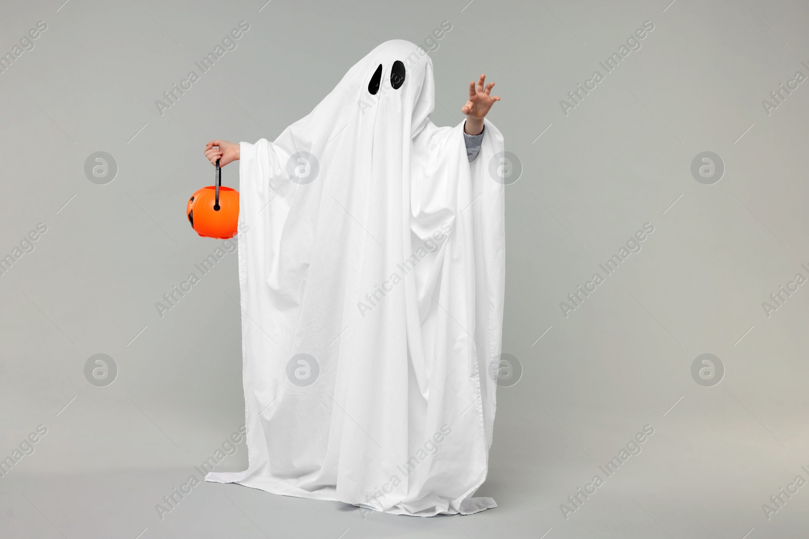 Photo of Child in white ghost costume holding pumpkin bucket on light grey background. Halloween celebration