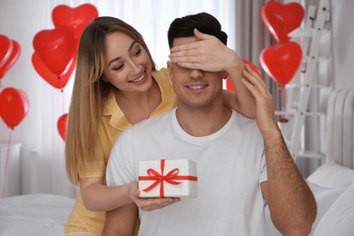 Photo of Woman presenting gift to her boyfriend in room decorated with heart shaped balloons. Valentine's day celebration