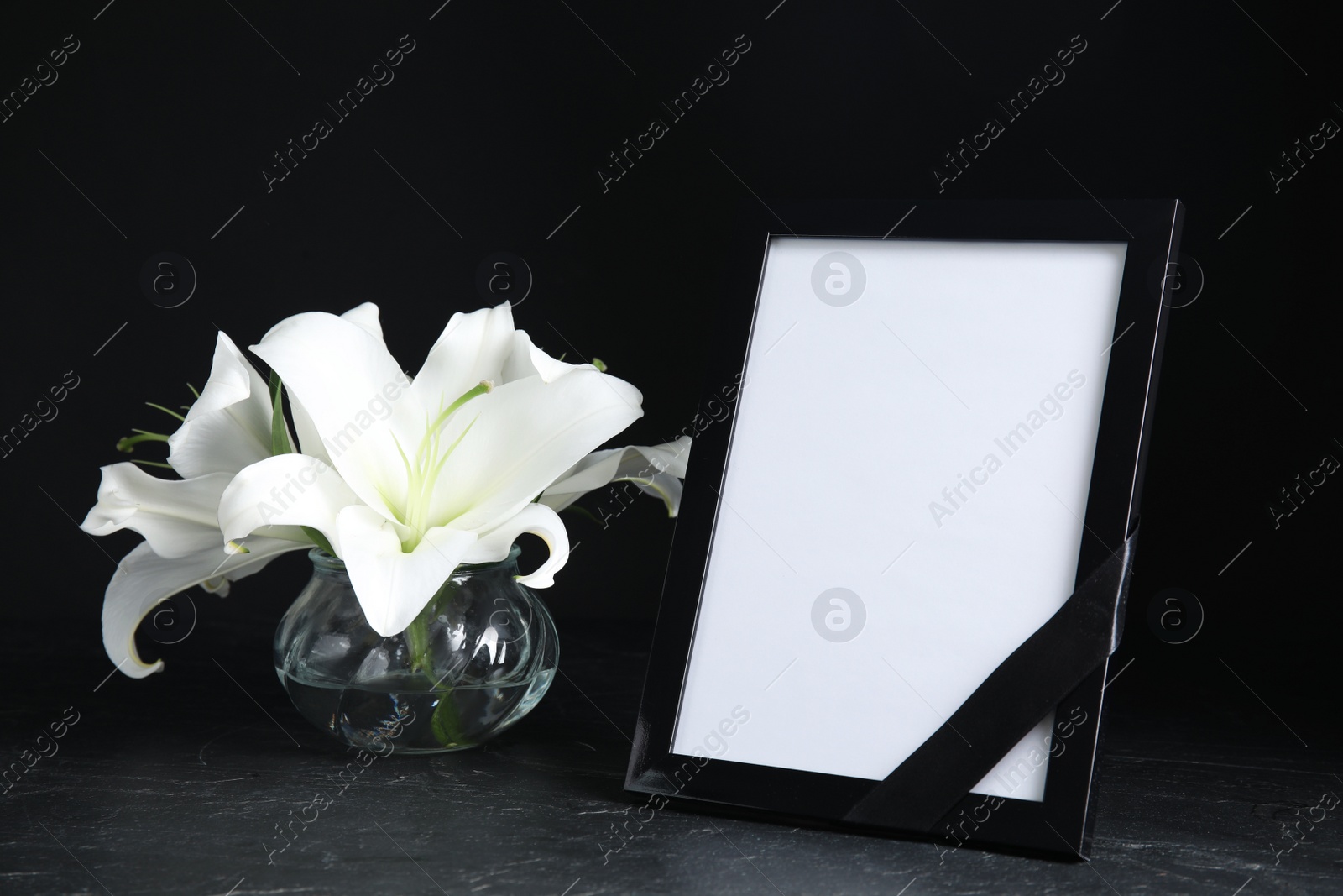 Photo of Funeral photo frame with ribbon and white lilies on black table against dark background. Space for design