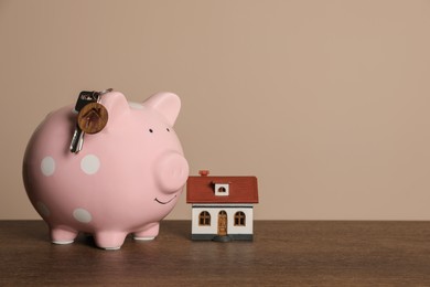 Photo of Piggy bank, keys and house model on wooden table against beige background. Space for text