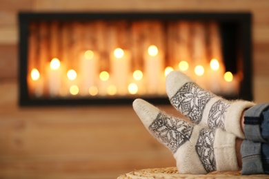 Photo of Woman wearing woolen socks relaxing on pouf against blurred background, closeup. Winter atmosphere
