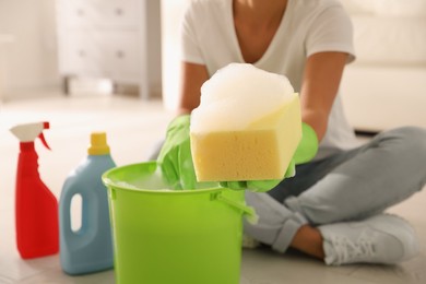 Photo of Woman holding sponge with foam over bucket indoors, closeup. Cleaning supplies