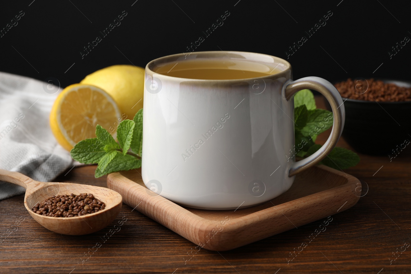 Photo of Cup of aromatic buckwheat tea, spoon with granules, lemon and mint on wooden table, closeup