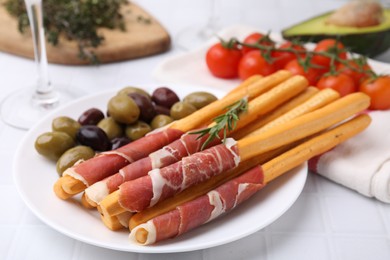 Photo of Plate of delicious grissini sticks with prosciutto and olives on white table, closeup