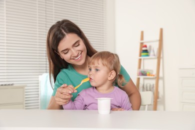 Mother feeding her cute little child with yogurt at white table in room