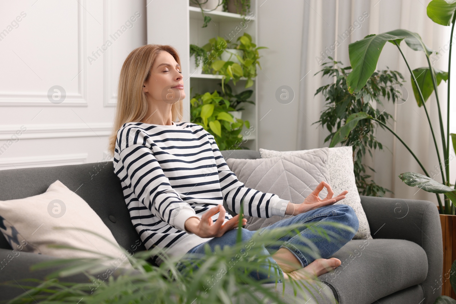 Photo of Woman meditating on sofa surrounded by beautiful potted houseplants at home